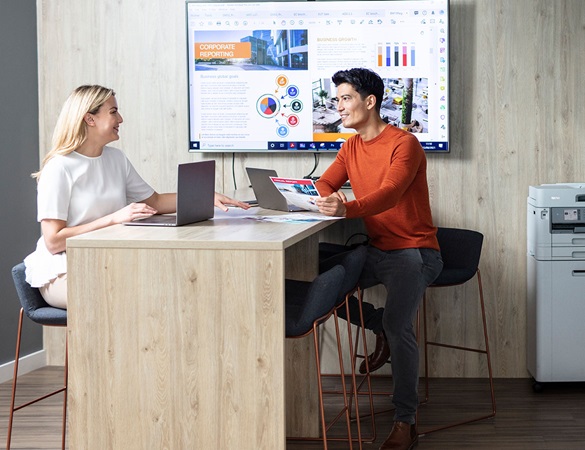 Mann og kvinne rundt et bord med TV på bakveggen og Brother skriver i bakgrunnen