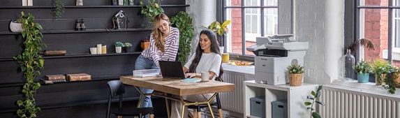 To kvinner sitter rundt skrivebordet i en  mindre bedrift, Brother-skriver, skap