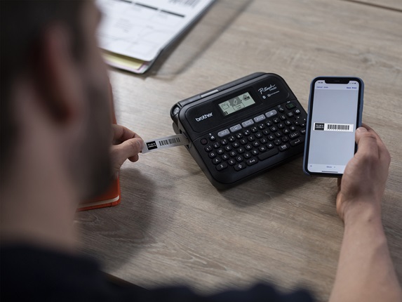 mann som sitter ved en pult med en p-touch maskin, holder en mobiltelefon i den ene hånden og en utskrevet etikett i den andre hånden