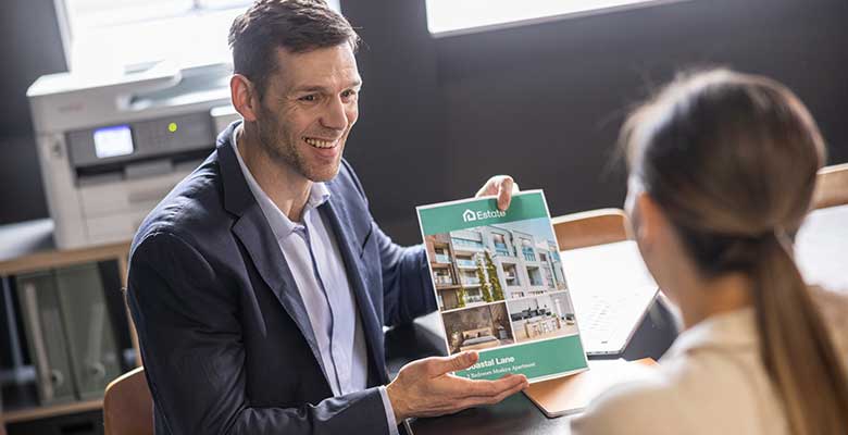 Mann holder opp farge A4-dokument som viser til kvinne, skriver, vindu