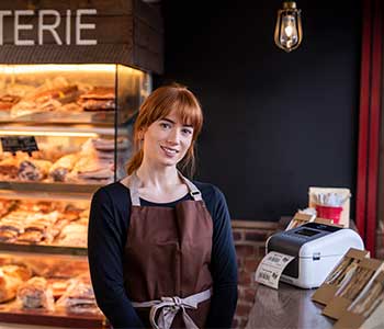 Kvinne med et brunt forkle i en kafe med en smørbrød og en TD-etiekttskriver ved siden av seg.