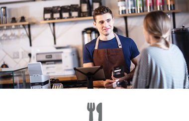 En ansatt i en kafe står bak disken og snakker med en kunde.  En Brother blekkskriver er plassert på en benk bak disken