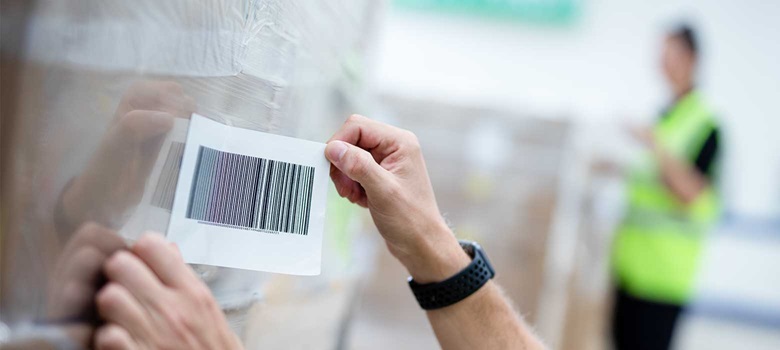 Etikett med strekkode festes på en pall med esker som har plast rundt seg på et lager
