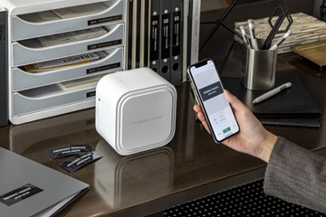 Brother Cube Pro PTP910BT merkemaskin og merkelapper på et bord. En hånd som holder en smarttelefon med en app i displayet. Permer og hyller merket med etiketter.